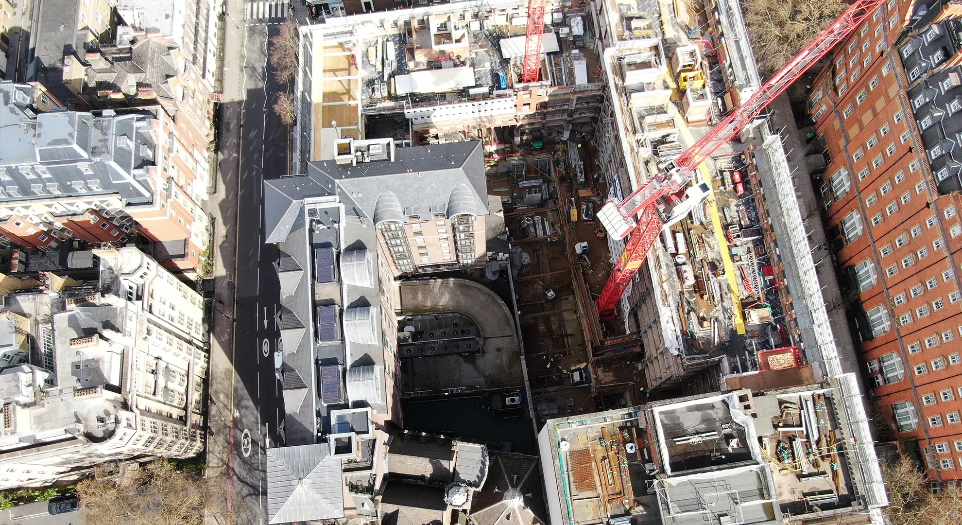 Top-down aerial drone view of an active urban construction site with a red tower crane and surrounding buildings.
