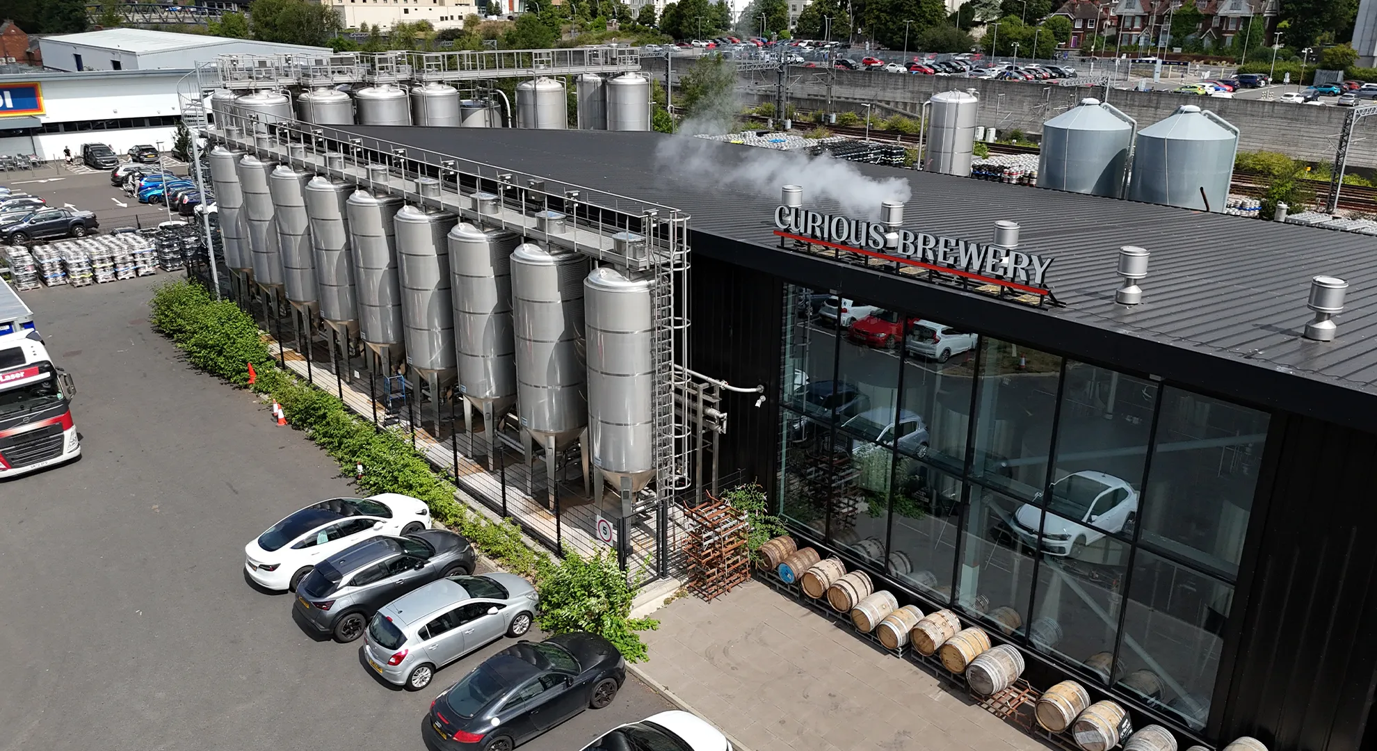 Elevated drone shot of Curious Brewery showing tall, stainless-steel cylindrical tanks lined up along a modern black building. Steam rises from a rooftop vent near the brewery logo, while wooden barrels line the pavement outside a large glass-front facade.