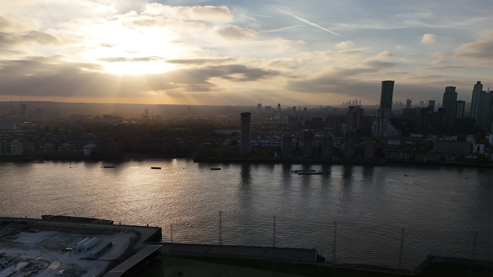 Wide aerial drone view looking across the River Thames toward the London skyline with sun rays breaking through clouds.