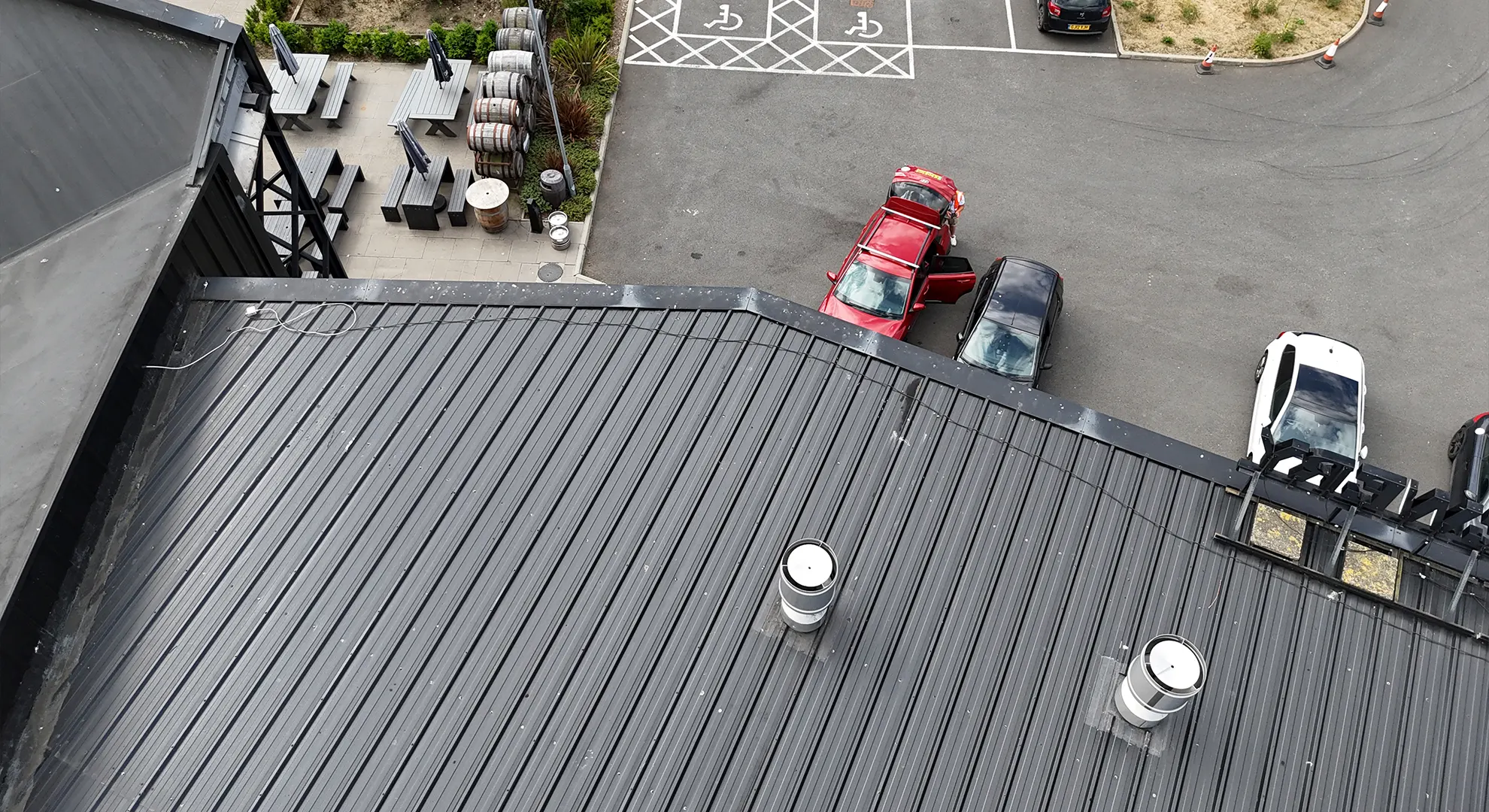 High-angle drone view of a dark corrugated metal roof overlooking a parking lot and outdoor patio area