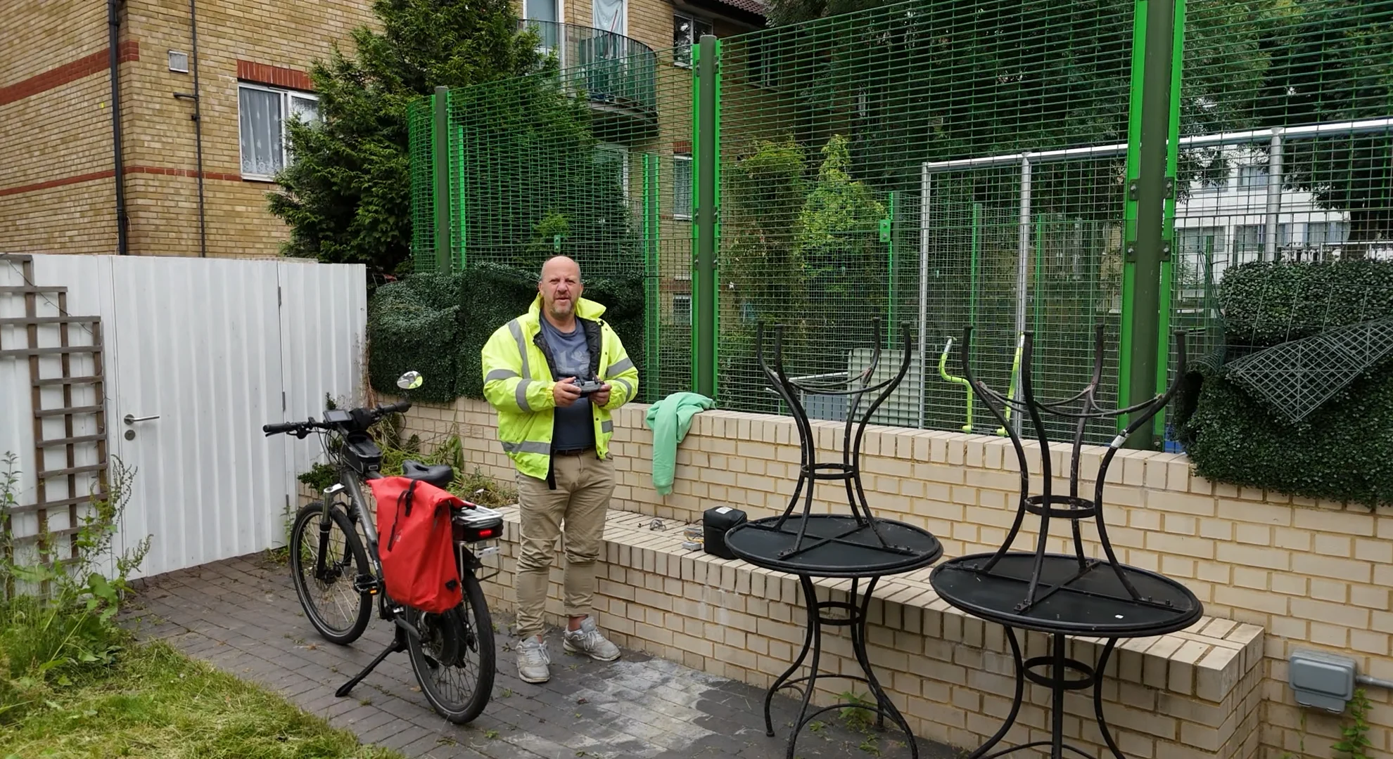 Drone camera shot as the drone takes off of man (Bob McGowan) in a high-visibility jacket holding a remote control next to a bicycle in an outdoor courtyard.