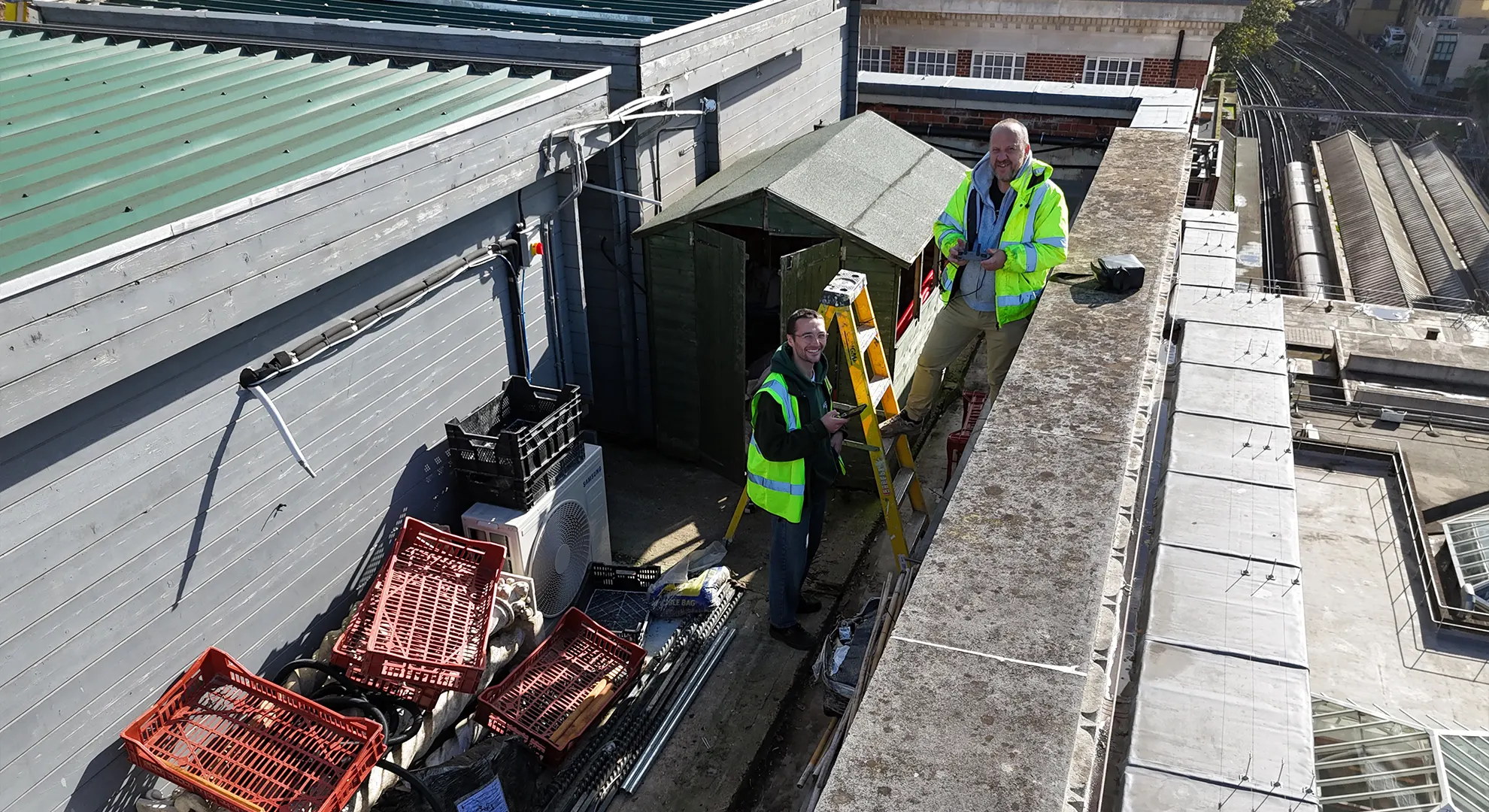 Two men in high-visibility vests on a rooftop, one holding a remote control, overlooking train tracks