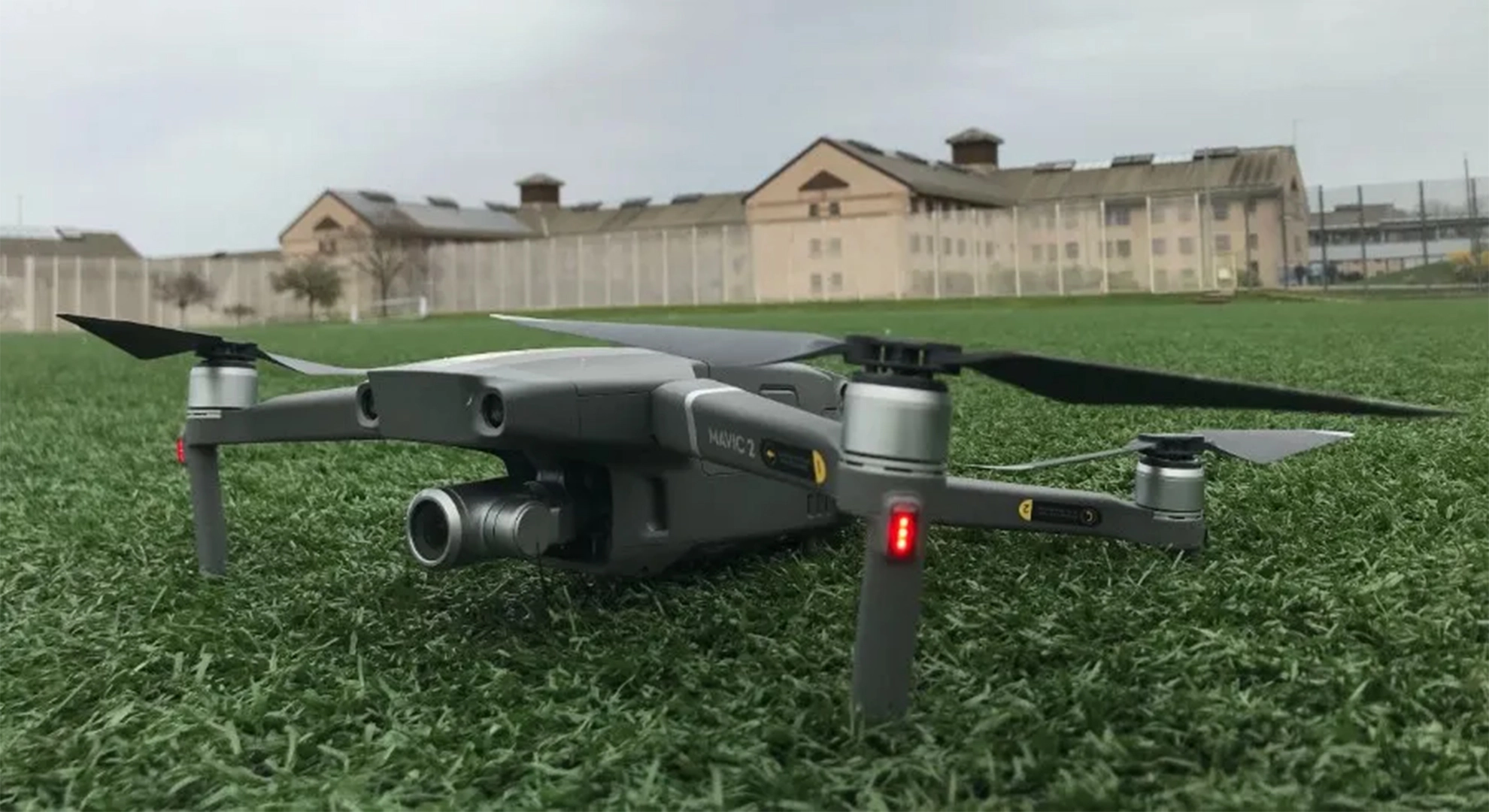 A DJI Mavic 2 drone on a green field with a large prison building and security fencing in the background