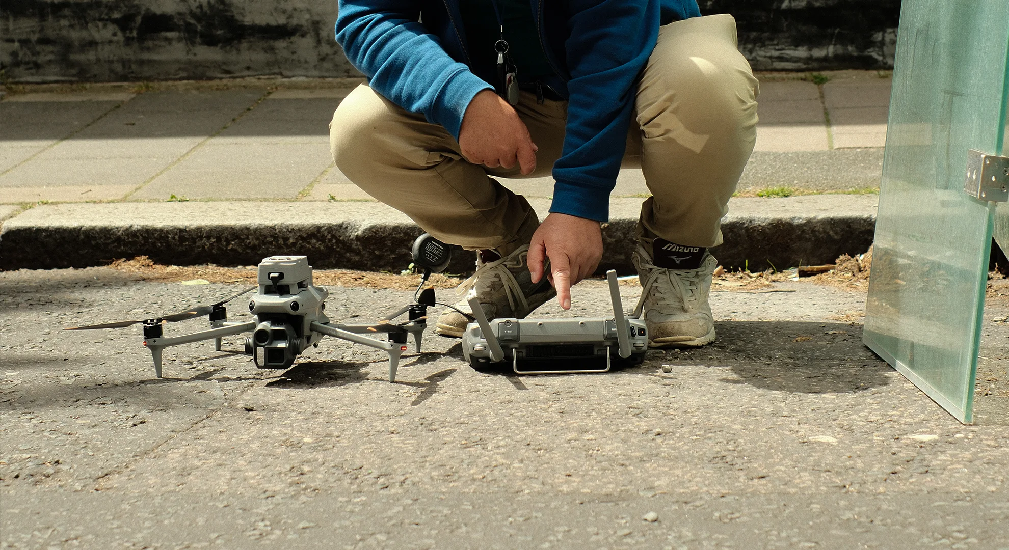 Helidrone Pilot Bob crouches on pavement, pointing at a drone touchscreen remote controller next to a grey quadcopter drone