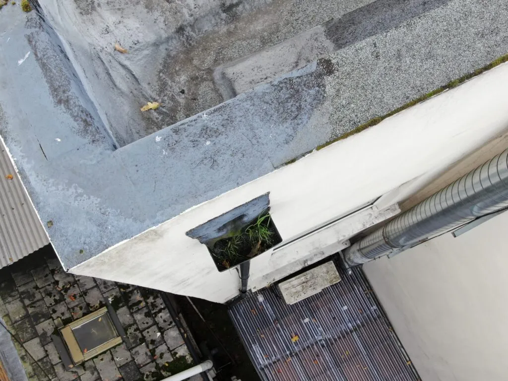 Close-up aerial drone inspection view of a flat roof corner showing a clogged gutter hopper head with growing weeds.