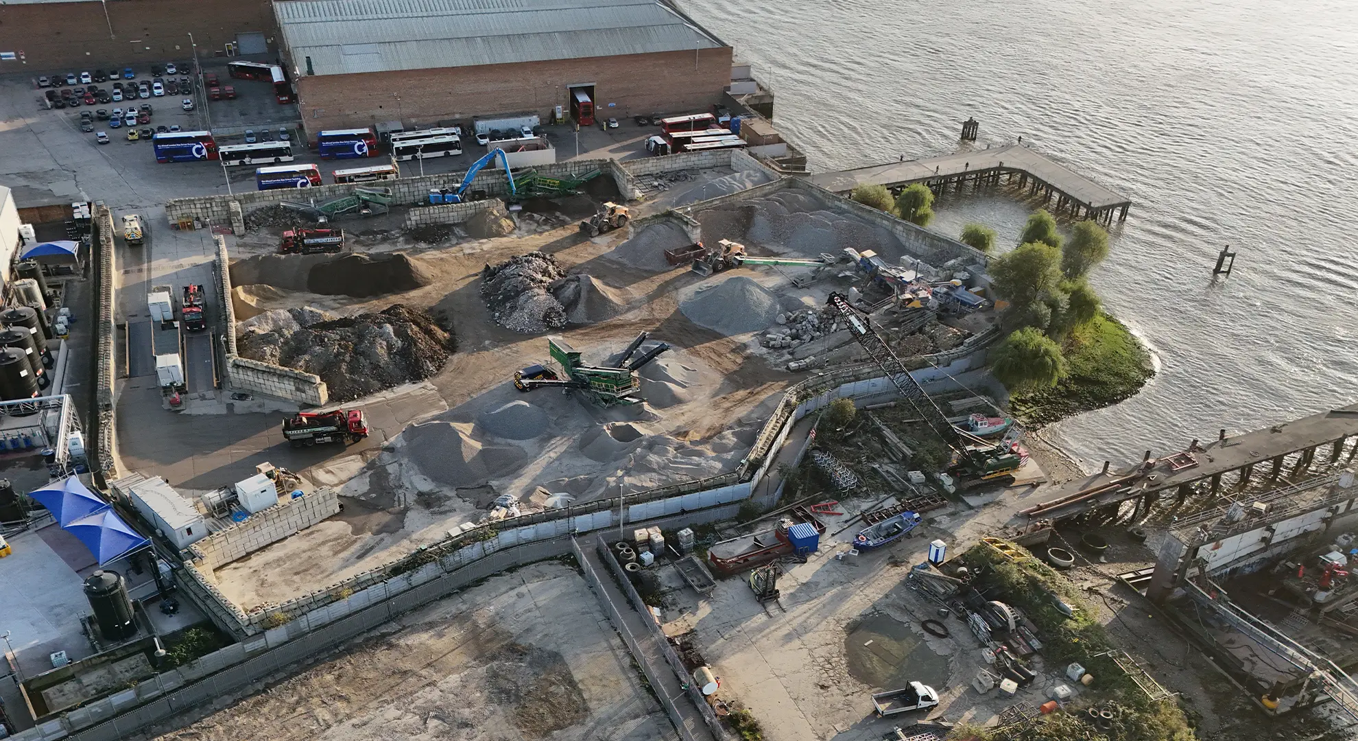 Aerial view of a riverside industrial site with gravel piles, excavators, a crane, and a bus depot