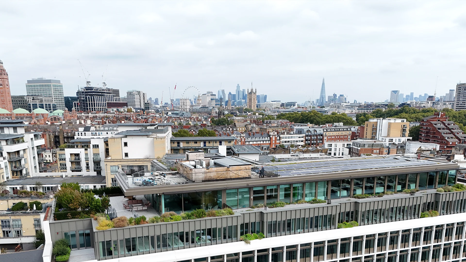 Elevated aerial drone view of a commercial building rooftop with solar panels and a terrace garden, overlooking the central London skyline.