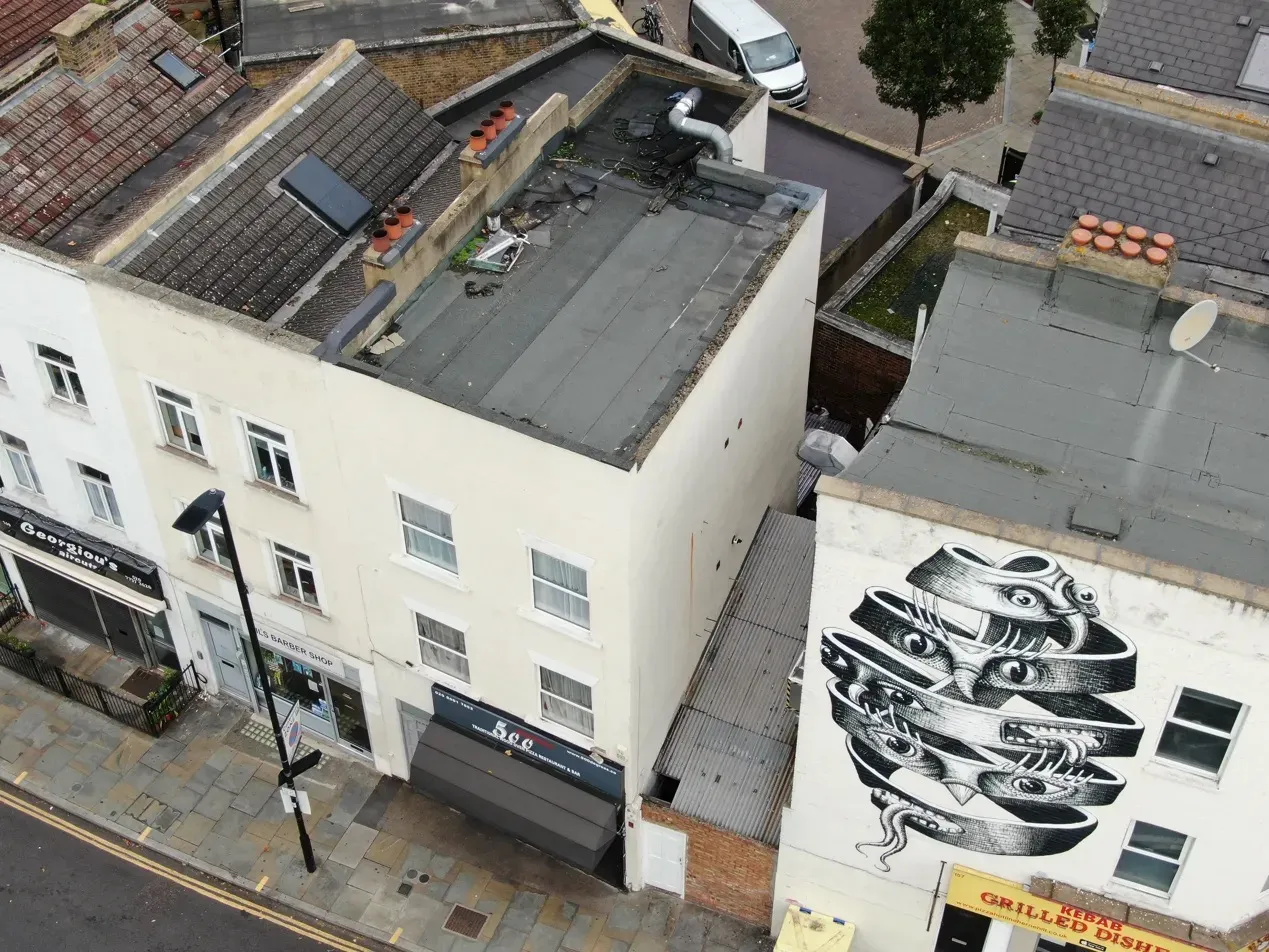 High-angle aerial drone view of commercial building rooftops showing flat roof condition, chimneys, and an exterior wall mural.