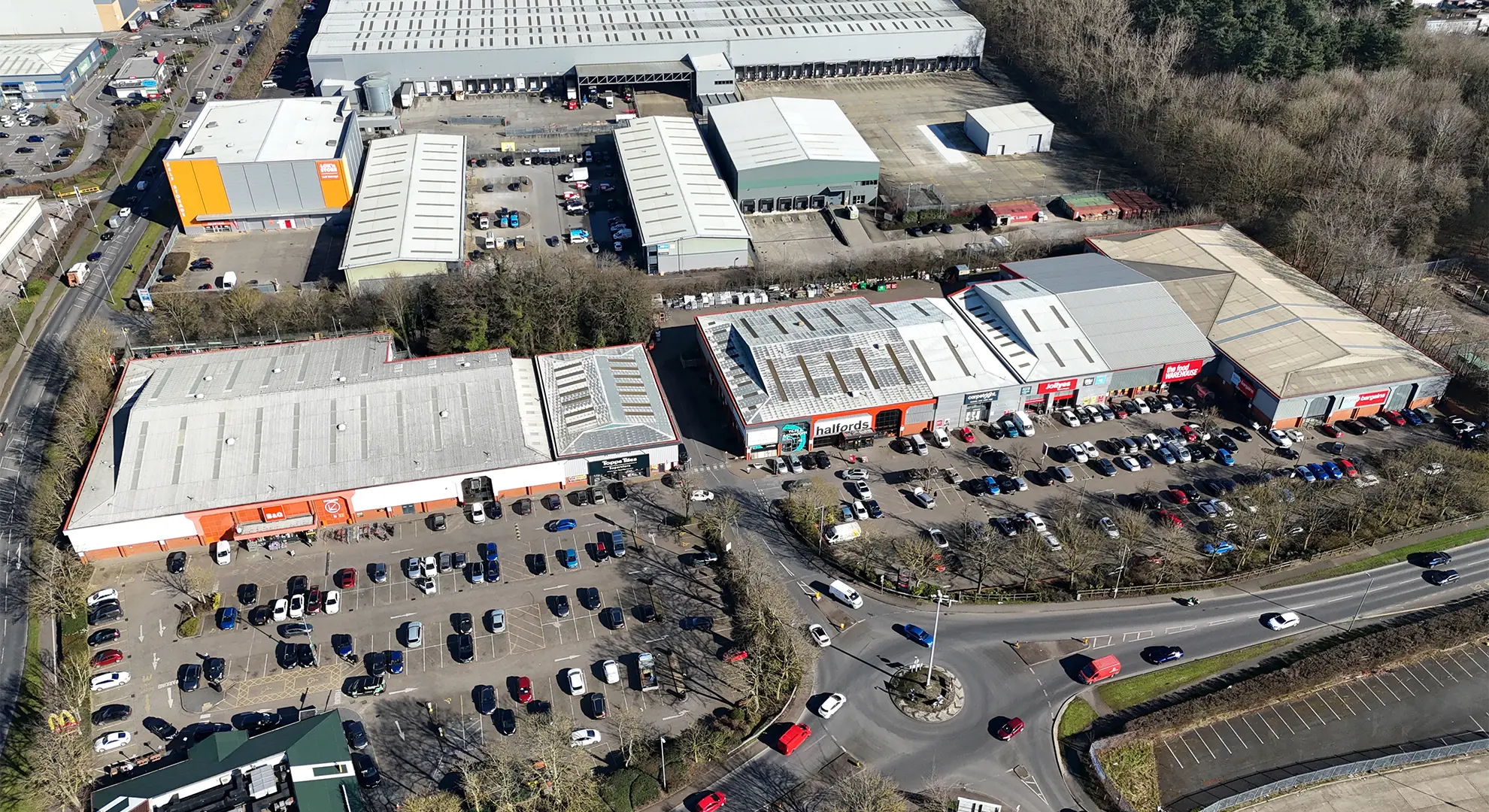 Wide aerial drone view overlooking a commercial retail park featuring warehouse rooftops, customer car parks, and an access roundabout.