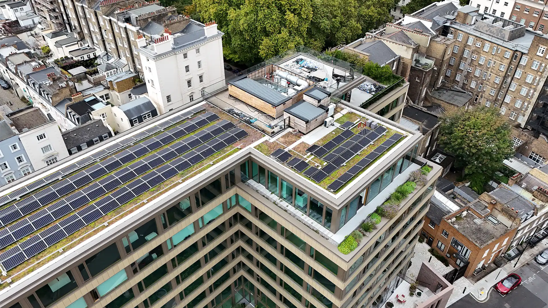 High-angle aerial drone view of a modern building rooftop fitted with extensive solar panel arrays, green roof sections, and surrounding urban streets.