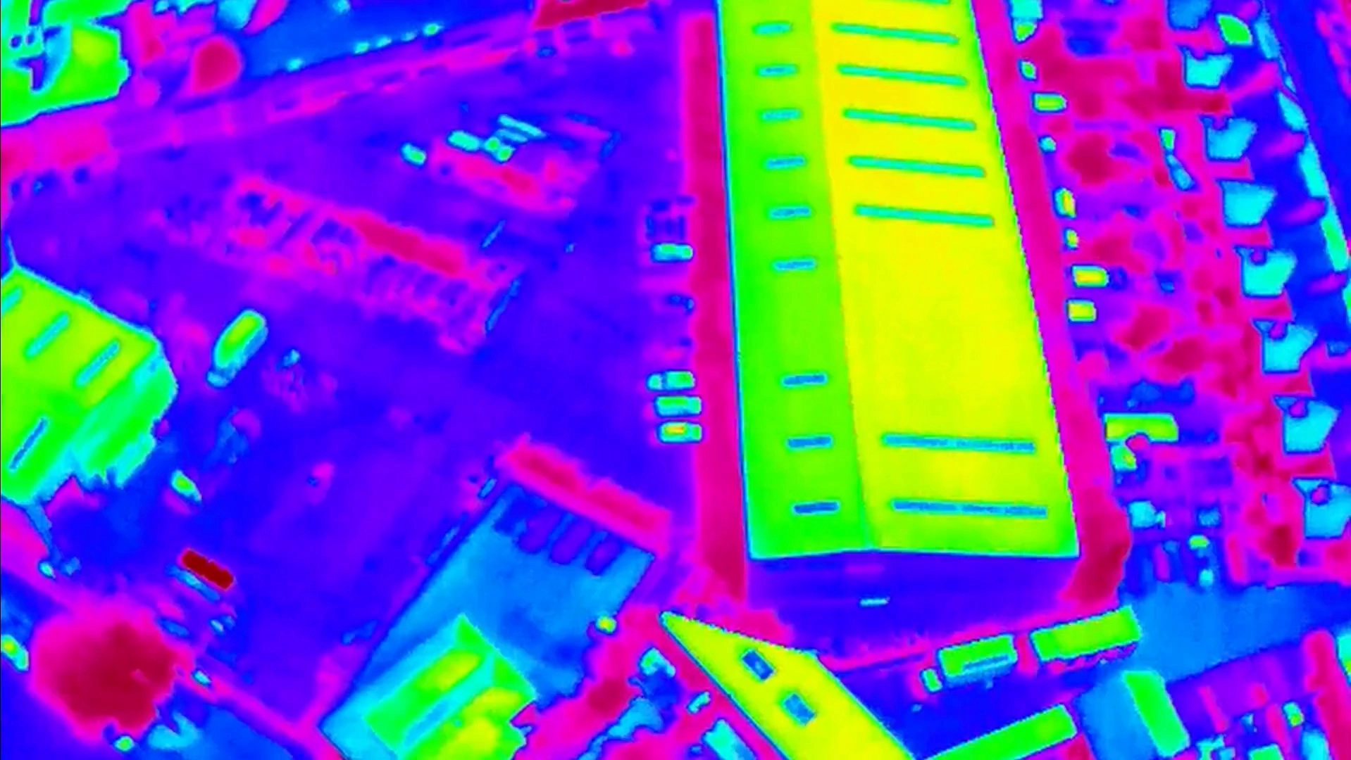 Aerial drone thermal imaging view of a large commercial building roof and surrounding site displayed in a high-contrast rainbow heat map palette.