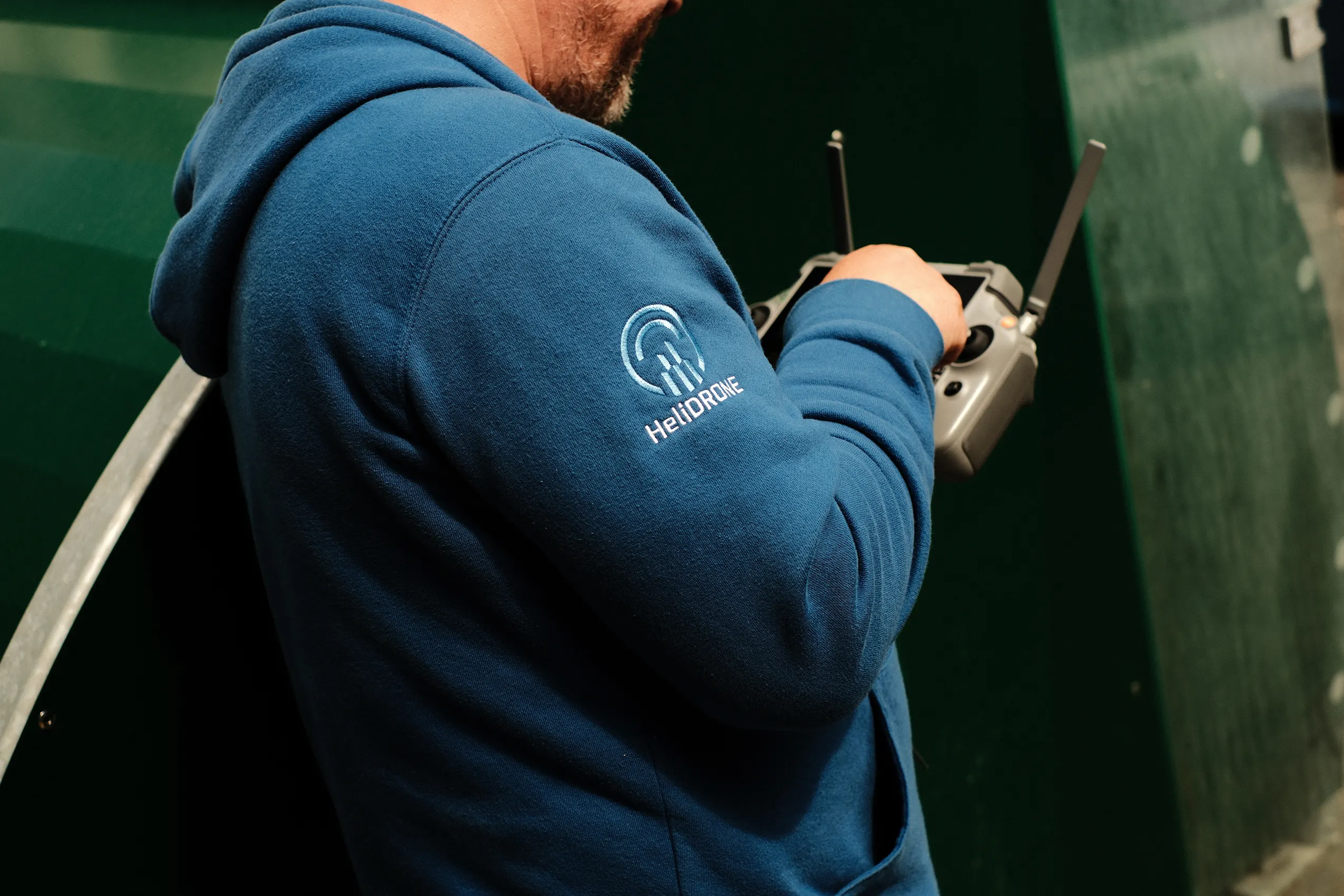 Side view of a drone operator wearing a blue HeliDRONE hoodie holding a remote controller.