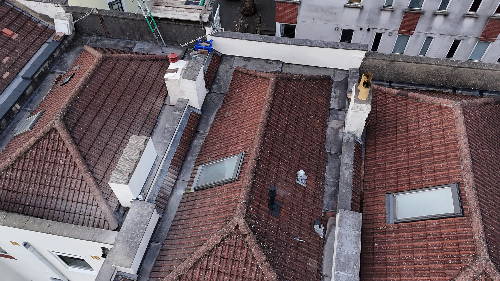 High-angle aerial drone roof inspection view showing multi-pitched red tile rooftops with chimney stacks, skylights, and lead valley gutters.