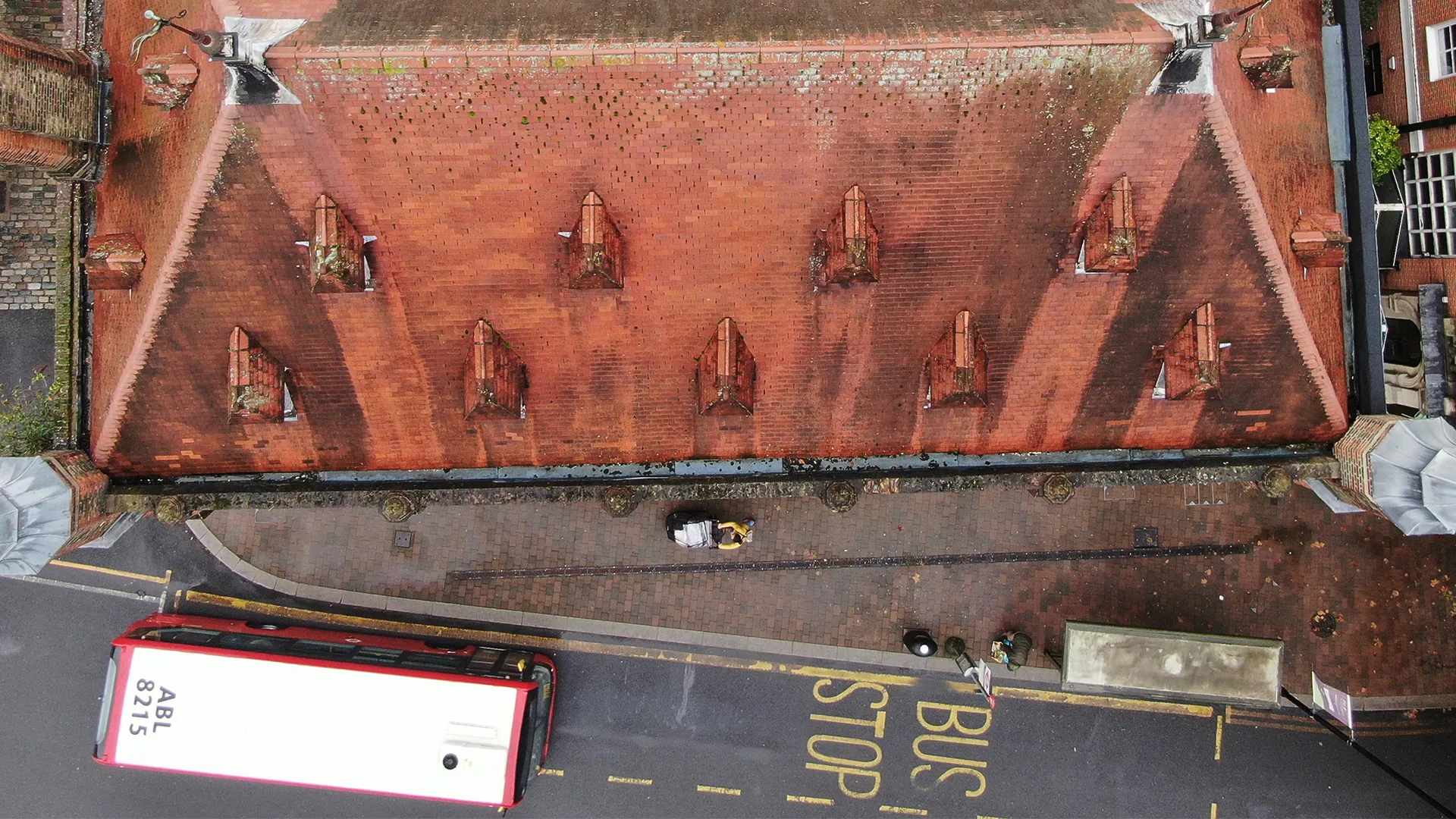 Top-down aerial drone inspection view of a weathered red clay tile roof with dormer vents above a street and bus stop with a red bus.