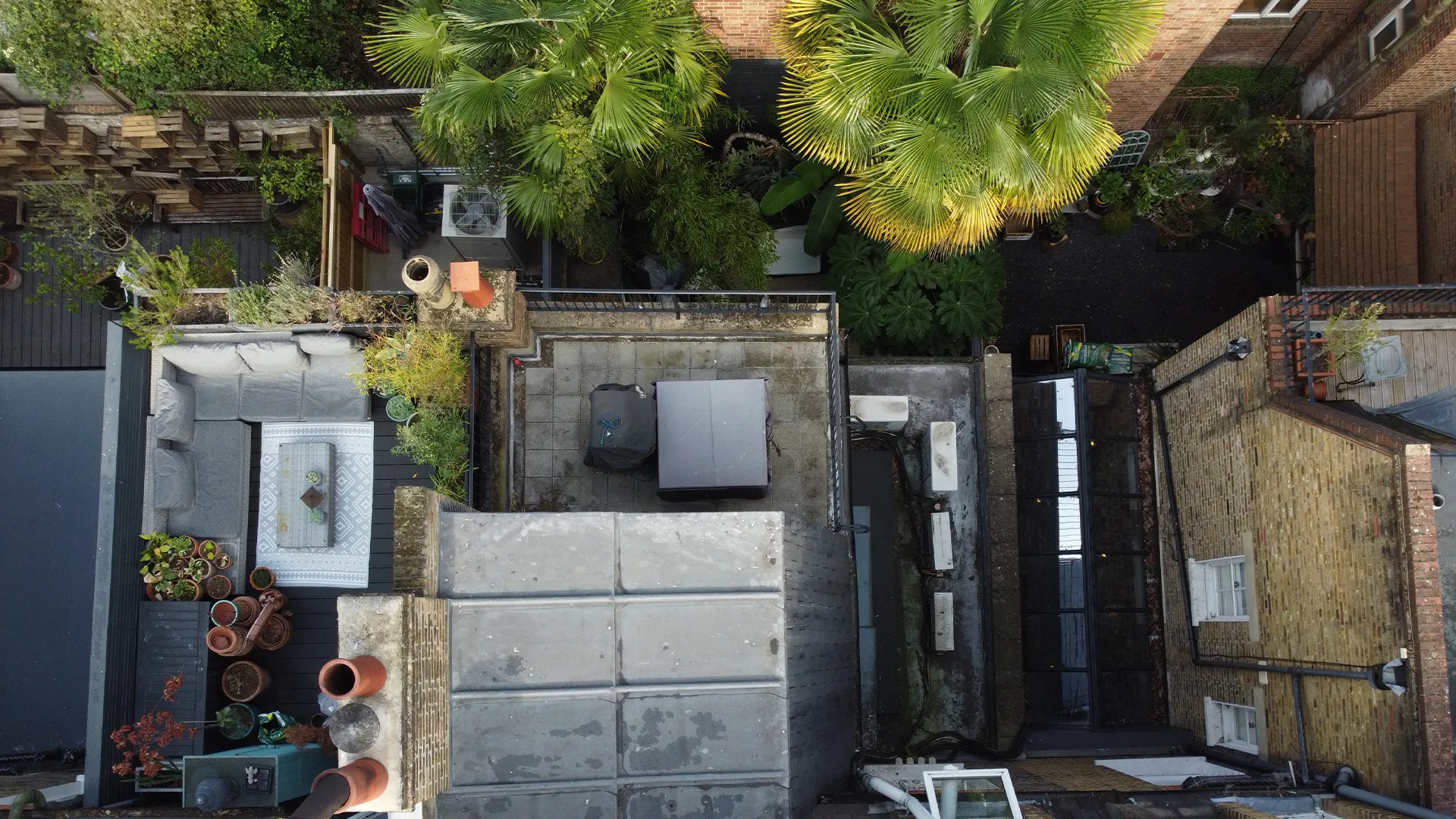 Overhead aerial drone inspection view of a residential building featuring a furnished rooftop terrace, chimney stacks, and a lush garden courtyard.