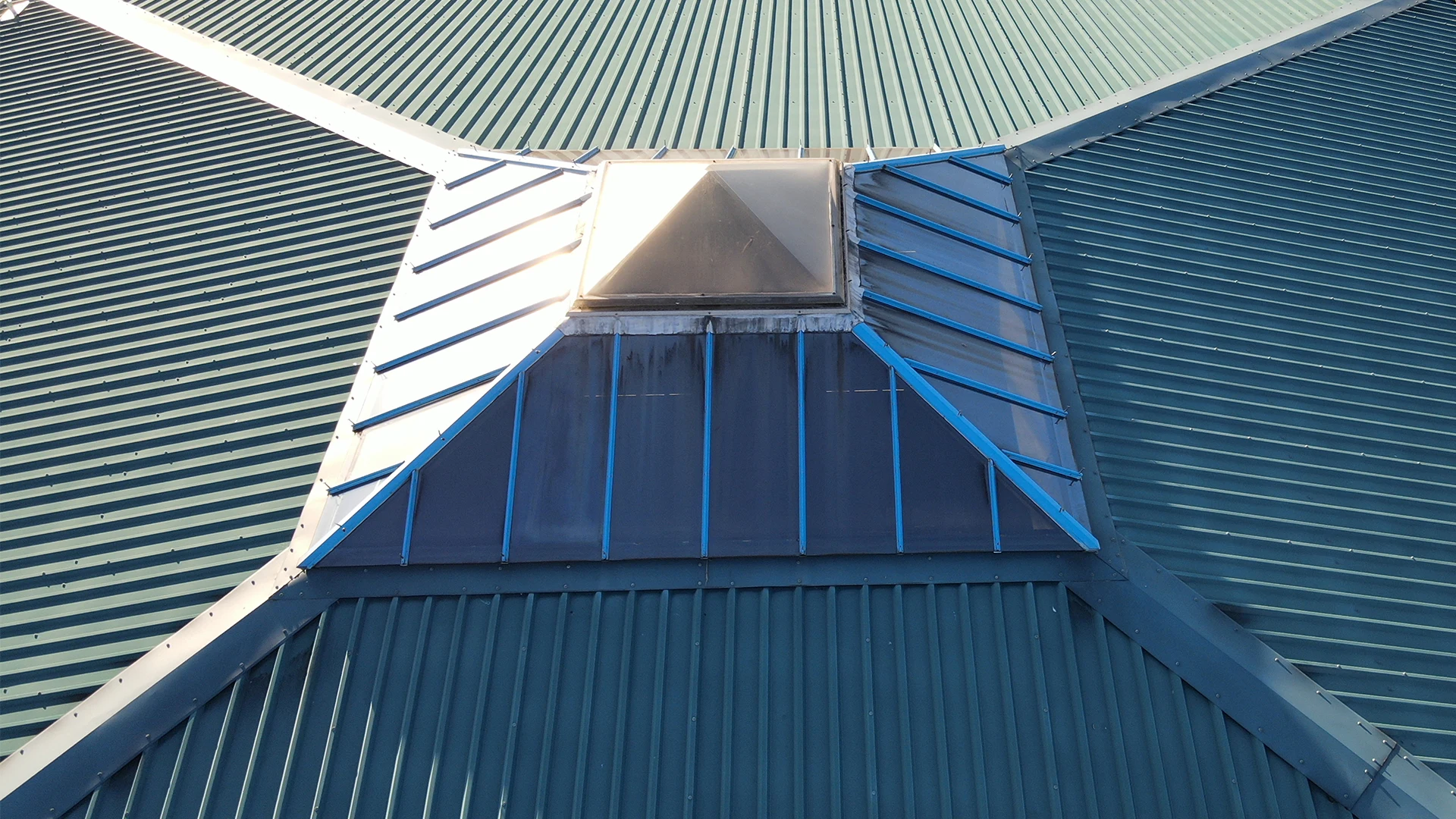 Close-up aerial drone roof inspection view of a green corrugated metal roof featuring a glazed skylight lantern reflecting sunlight.